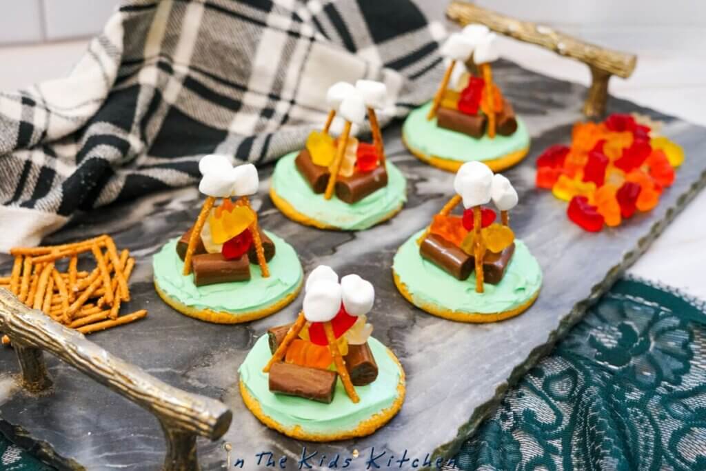gray platter displaying campfire sugar cookie with green icing, tootsie roll logs, gummy bear flames and pretzel smores sticks
